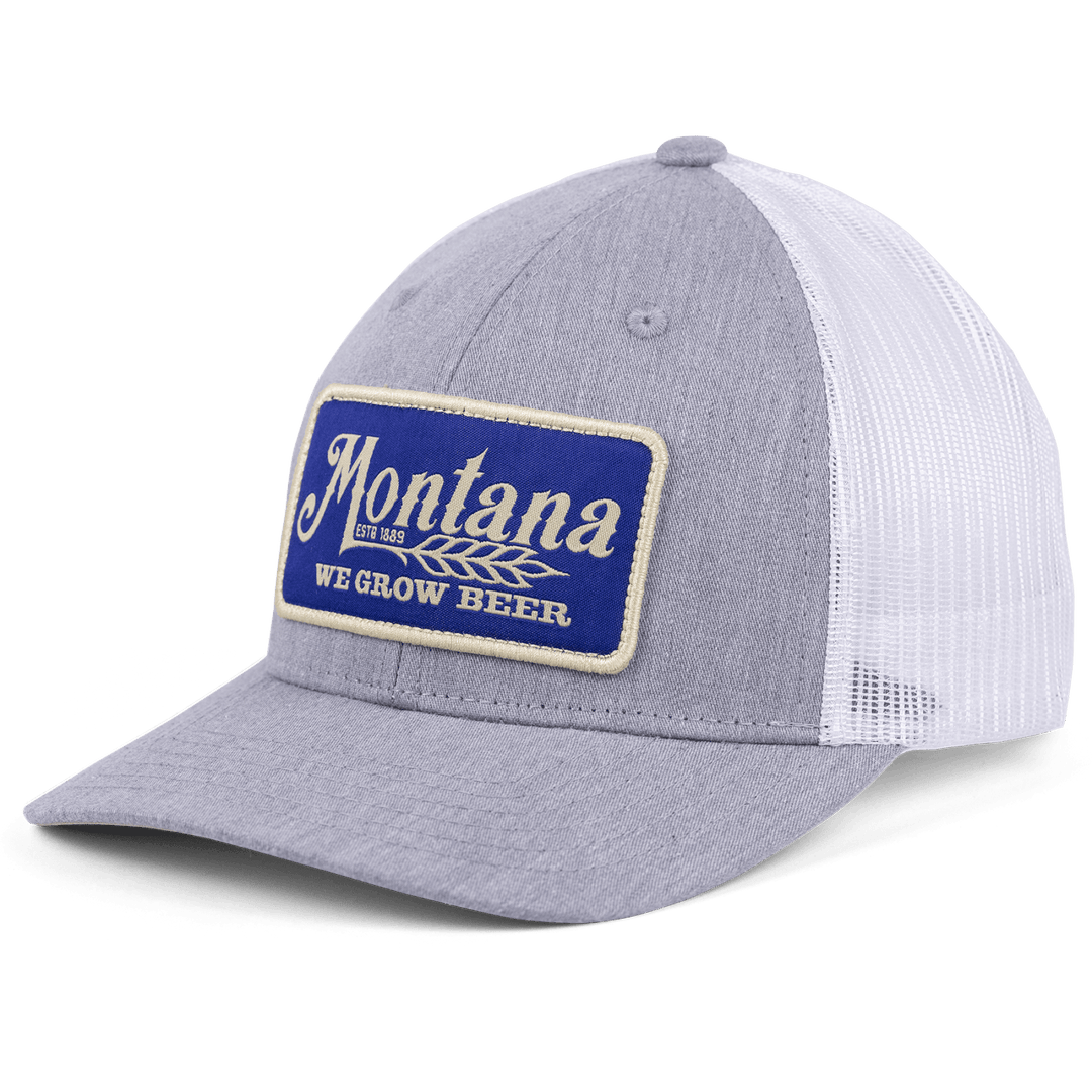 The Montana We Grow Beer Hat by Rural Cloth is a grey and white trucker style featuring a blue patch embroidered with "Montana, Est. 1889, WE GROW BEER" on the front. It boasts an adjustable snapback closure and a breathable mesh back for comfort.