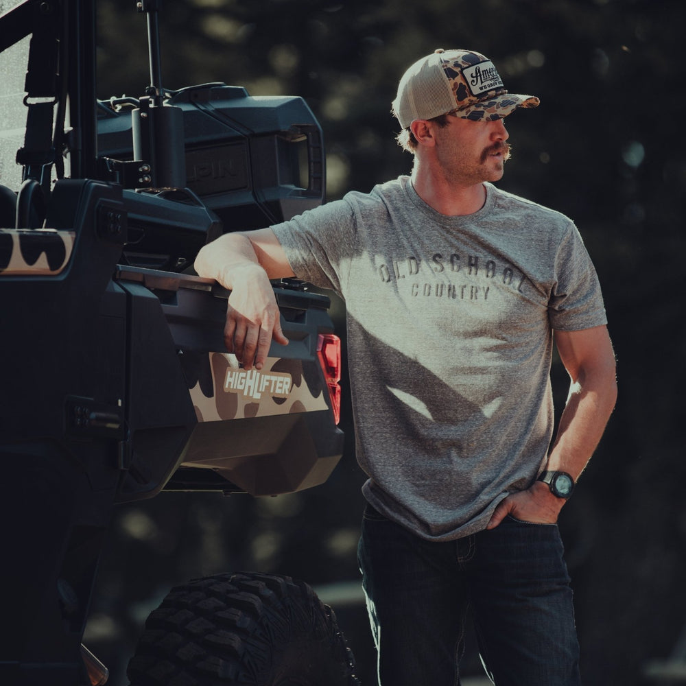 A person wearing an Old School Country - Heather Gray T-shirt from Rural Cloth and a camo cap stands next to an off-road vehicle. With one arm leaning on the vehicle, they gaze into the distance. The vehicle features a "High Lifter" logo, and the outdoor setting in the background is out of focus.
