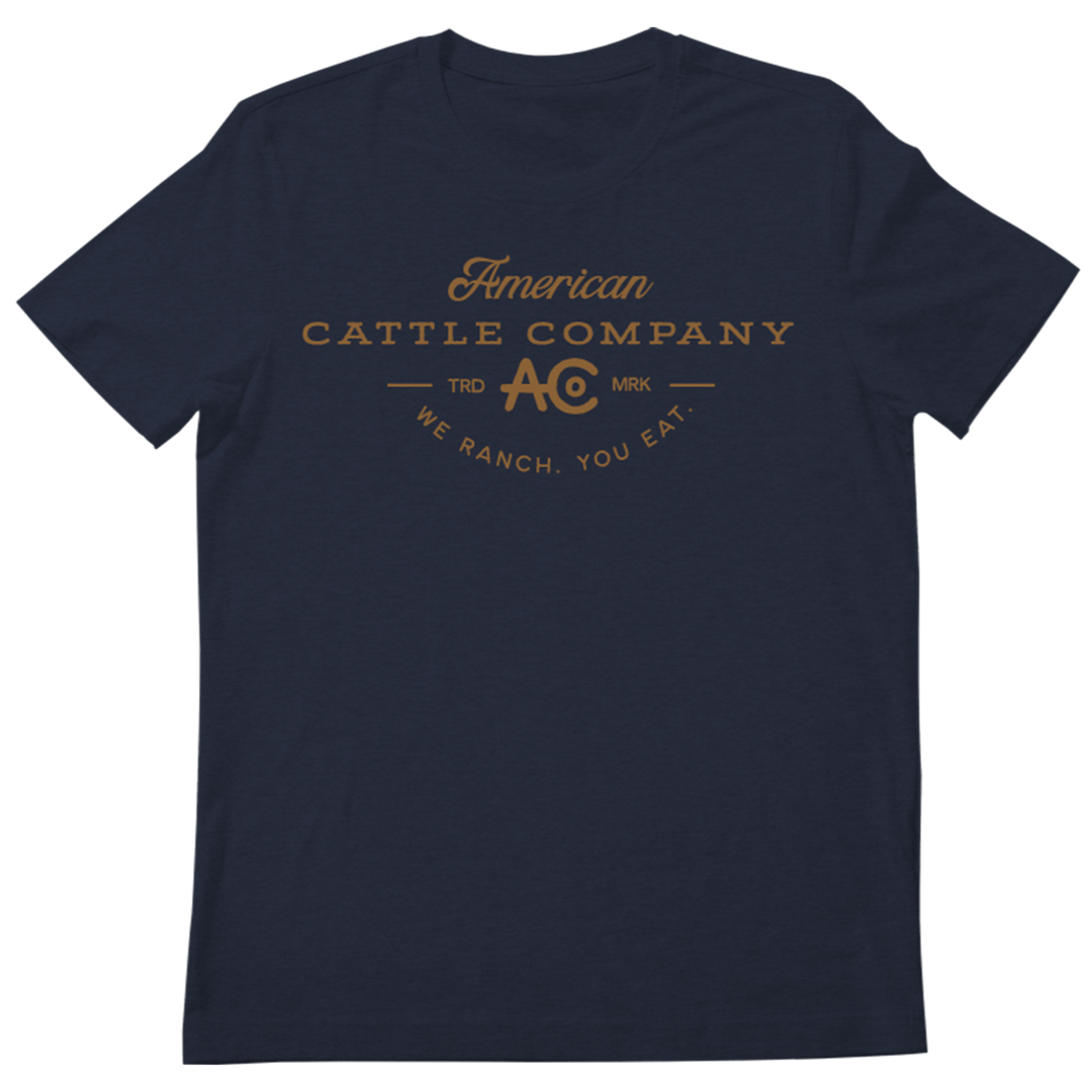 A heather navy Provider Tee by Rural Cloth features the text "American Cattle Company" printed in brown across the chest. Below it are the “AC” and “MRK” logos, followed by the slogan “We Ranch. You Eat.” This t-shirt has a simple, classic design with short sleeves and a round neck, celebrating cattle farming heritage.