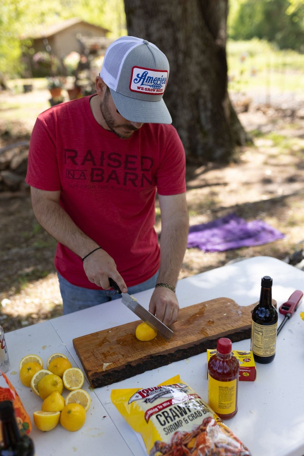 A man wearing a red "Raised In A Barn" vintage fitted shirt by Rural Cloth and a baseball cap is cutting a lemon on a wooden cutting board outdoors. Various ingredients, including Cajun seasoning and lemons, are on the table. There is a tree and greenery in the background.