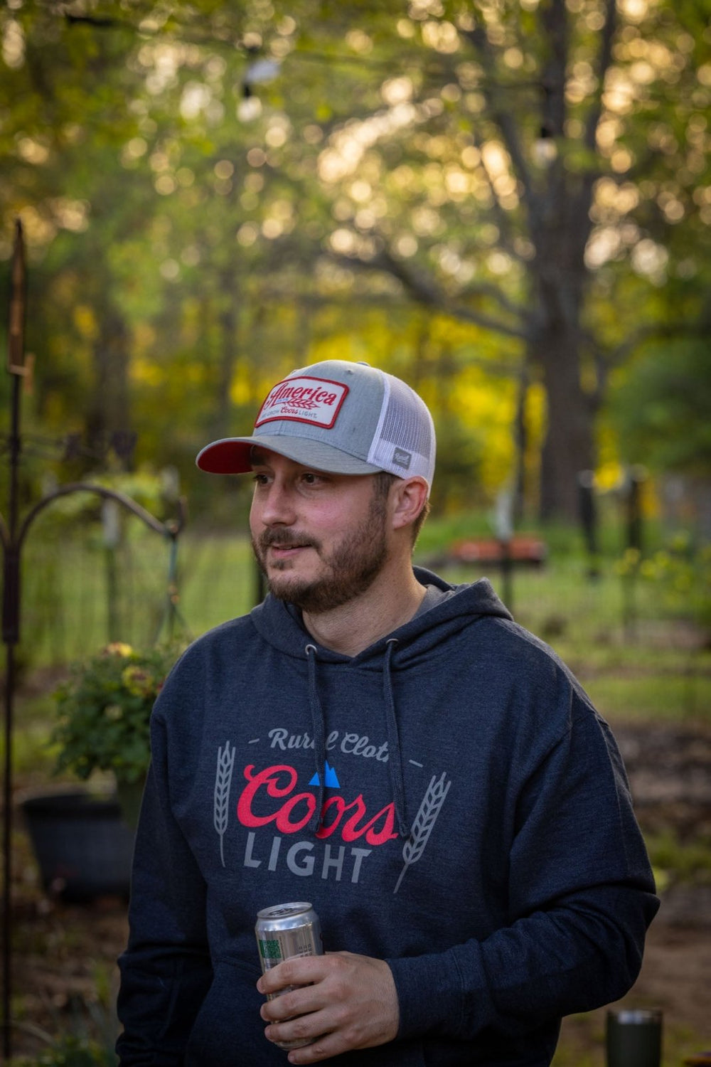 A man wearing a heather navy RC x CL Pullover by Rural Cloth, featuring the Coors Light logo, and a red and white cap stands outdoors. He holds an American beer can in his left hand as the background shows a garden with greenery and trees.