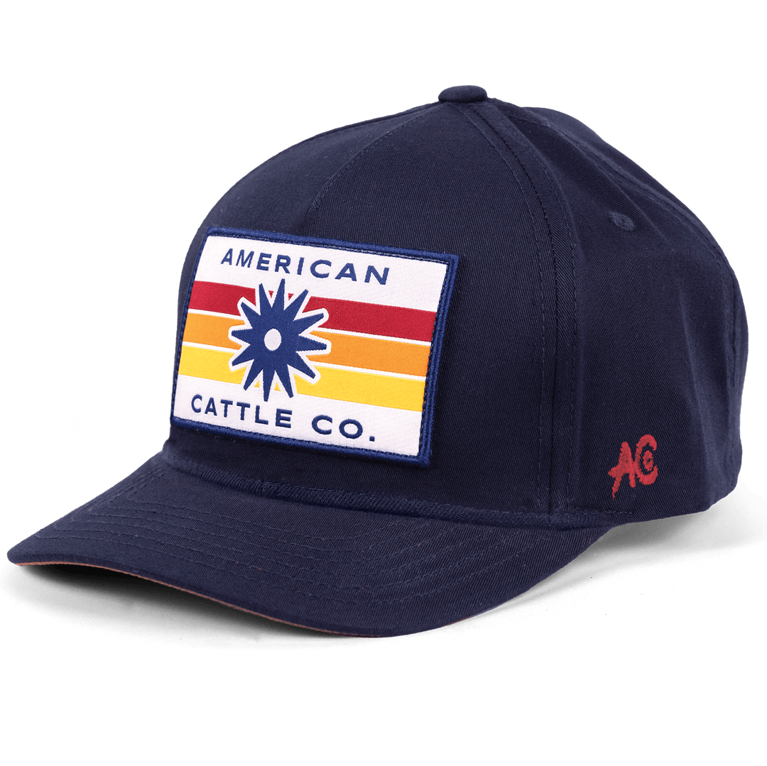 The Retro Spur Hat by Rural Cloth is a navy blue baseball cap featuring a rectangular patch on the front with the text "American Cattle Co." in blue and white, set against red, yellow, and orange horizontal stripes. Embracing western culture, this adjustable snapback closure hat has the initials "AC" in red on its left side.