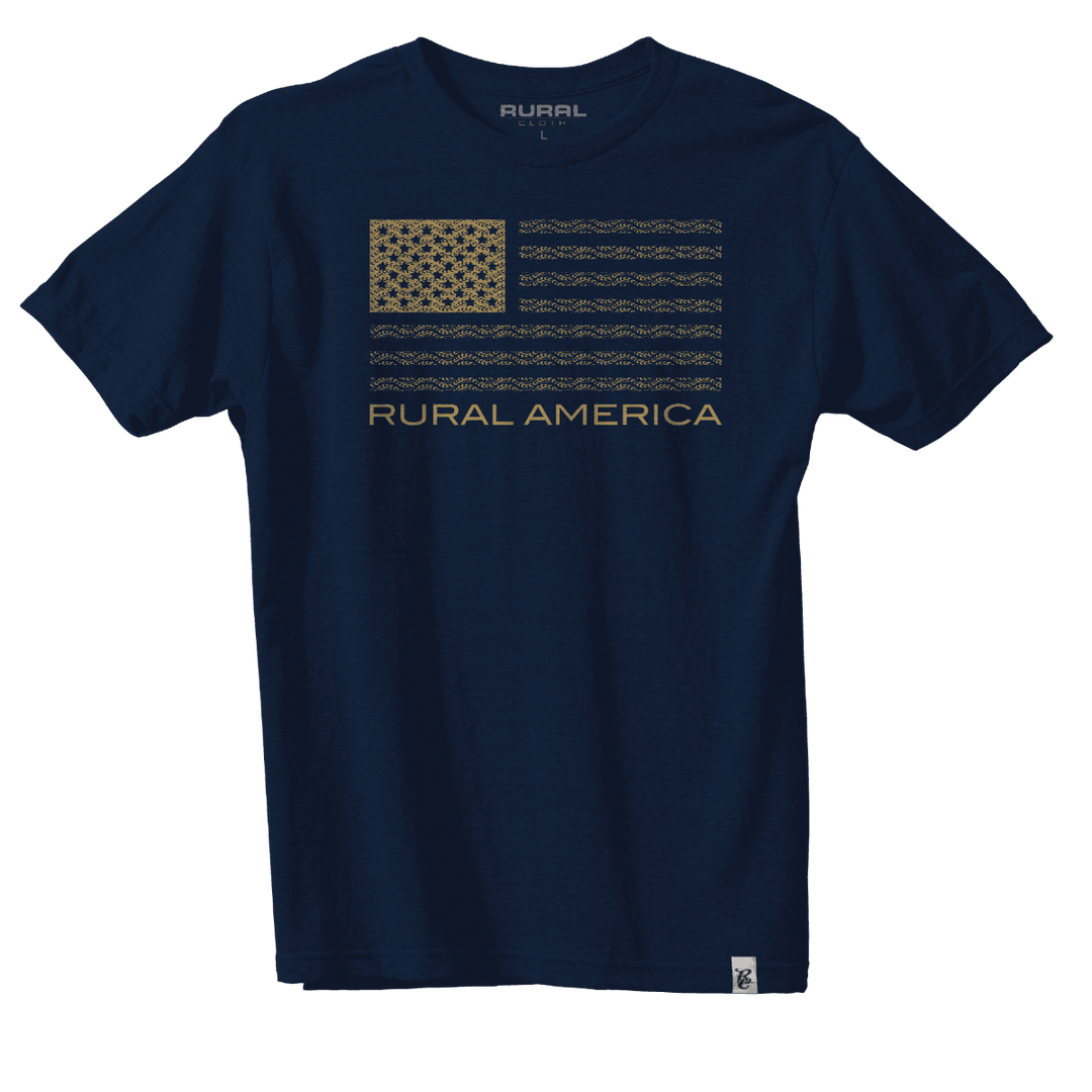 The "Rural America Wheat Flag - Navy Blue" T-shirt by Rural Cloth features a stylized American flag in shades of tan and olive green on the chest, along with the text "RURAL AMERICA" in tan below. This durable poly-blend shirt is showcased against a plain white background.
