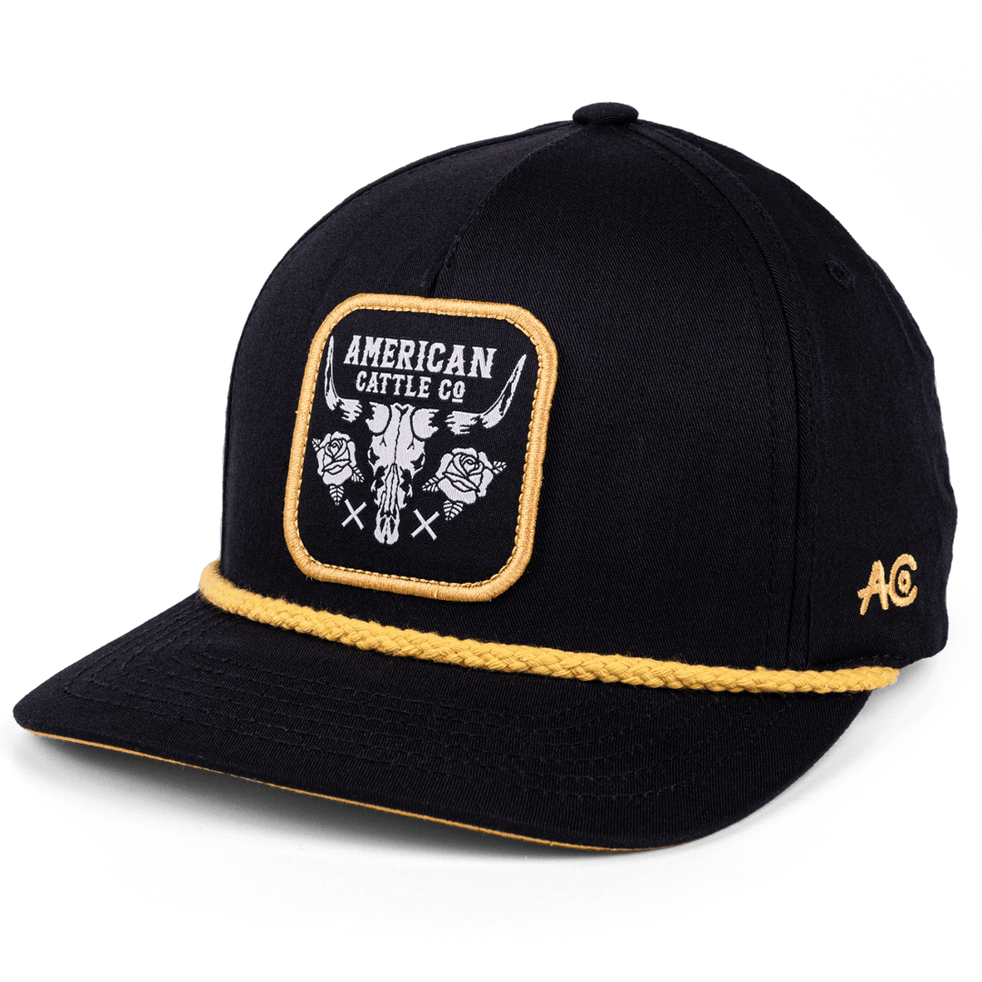 A black adjustable Skull & Roses Hat by Rural Cloth, featuring yellow trim and a front patch that reads "AMERICAN CATTLE CO" with a skull and roses design. The letters "AC" are embroidered in yellow on the side of this western-style snapback.