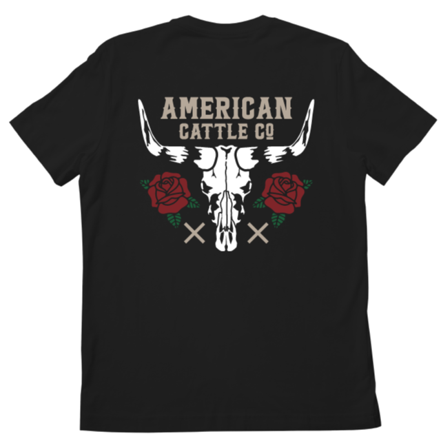 The image displays the back of a Skull & Roses Tee-Black by Rural Cloth, featuring a large bull skull flanked by two red roses and green leaves. Above the skull, bold text reads "AMERICAN CATTLE CO," making this tee a striking statement piece.