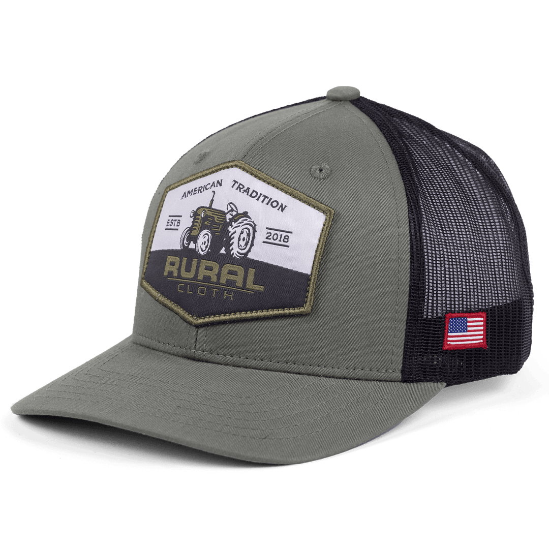 The Tractor Hat from Rural Cloth, featuring a gray and black design with a mesh back, includes an embroidered patch showcasing a tractor along with the text "American Tradition: Estd 2018" and "Rural Cloth." An American flag patch is also positioned on the side. The adjustable snapback closure guarantees a perfect fit for any proud American farmer.