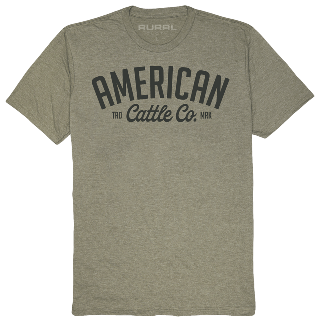 The Trademark Tee is a light olive-green T-shirt made from premium weight fabric with "American Cattle Co." printed in large, bold letters on the front. The word "RURAL" is printed on the inside neckline. This T-shirt from Rural Cloth boasts a simple, clean design with short sleeves and a crew neck.