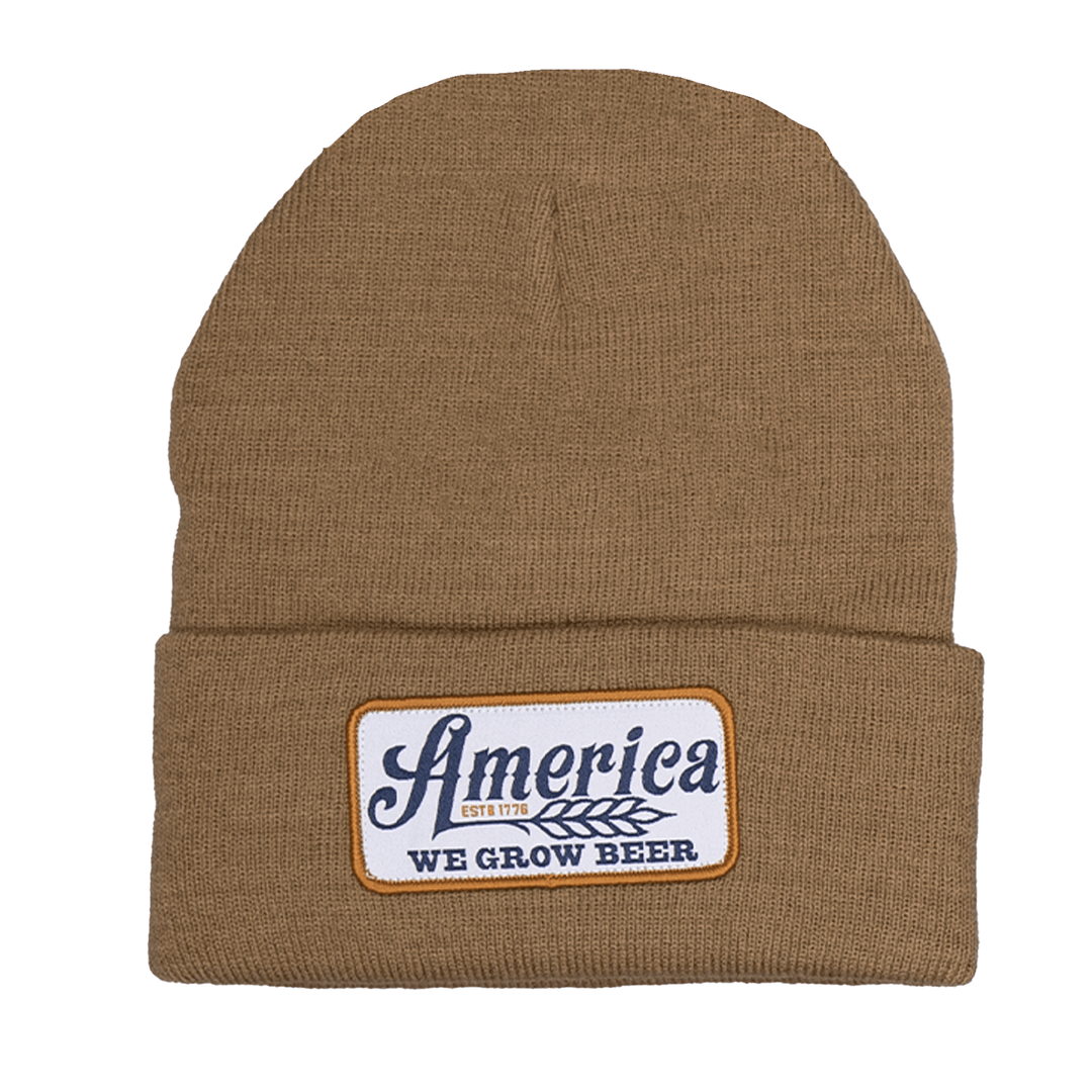 The We Grow Beer Beanie by Rural Cloth is a brown knit hat featuring a patch on the front that reads "America EST. 1776" in blue text and "WE GROW BEER" in white text, bordered by yellow. This beanie pays tribute to American workers.