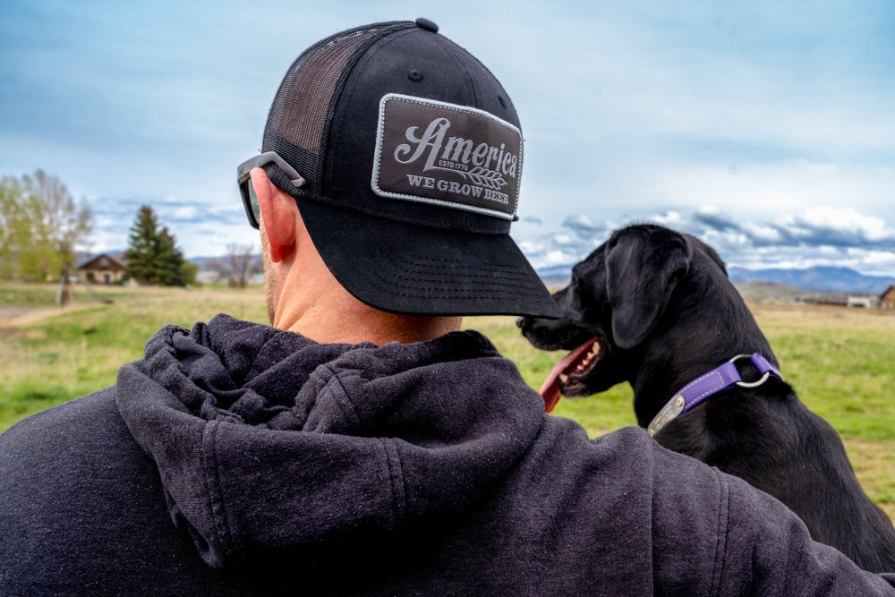 A person wearing a black hoodie and a Rural Cloth's We Grow Beer Hat-Black, which features an adjustable snapback closure, is standing outside in a field, holding a black dog with a purple collar. The background showcases a grassy area with trees and a partly cloudy sky.