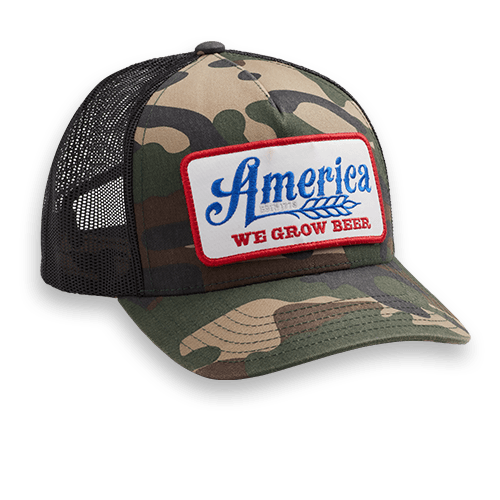 Introducing the We Grow Beer Hat-Camo from Rural Cloth: a camouflage baseball cap with a black mesh back. The front showcases a rectangular patch, featuring blue text that reads "America" and red text below that says "WE GROW BEER." This hat, edged in red and adorned with a wheat graphic, pays homage to hard-working Americans.