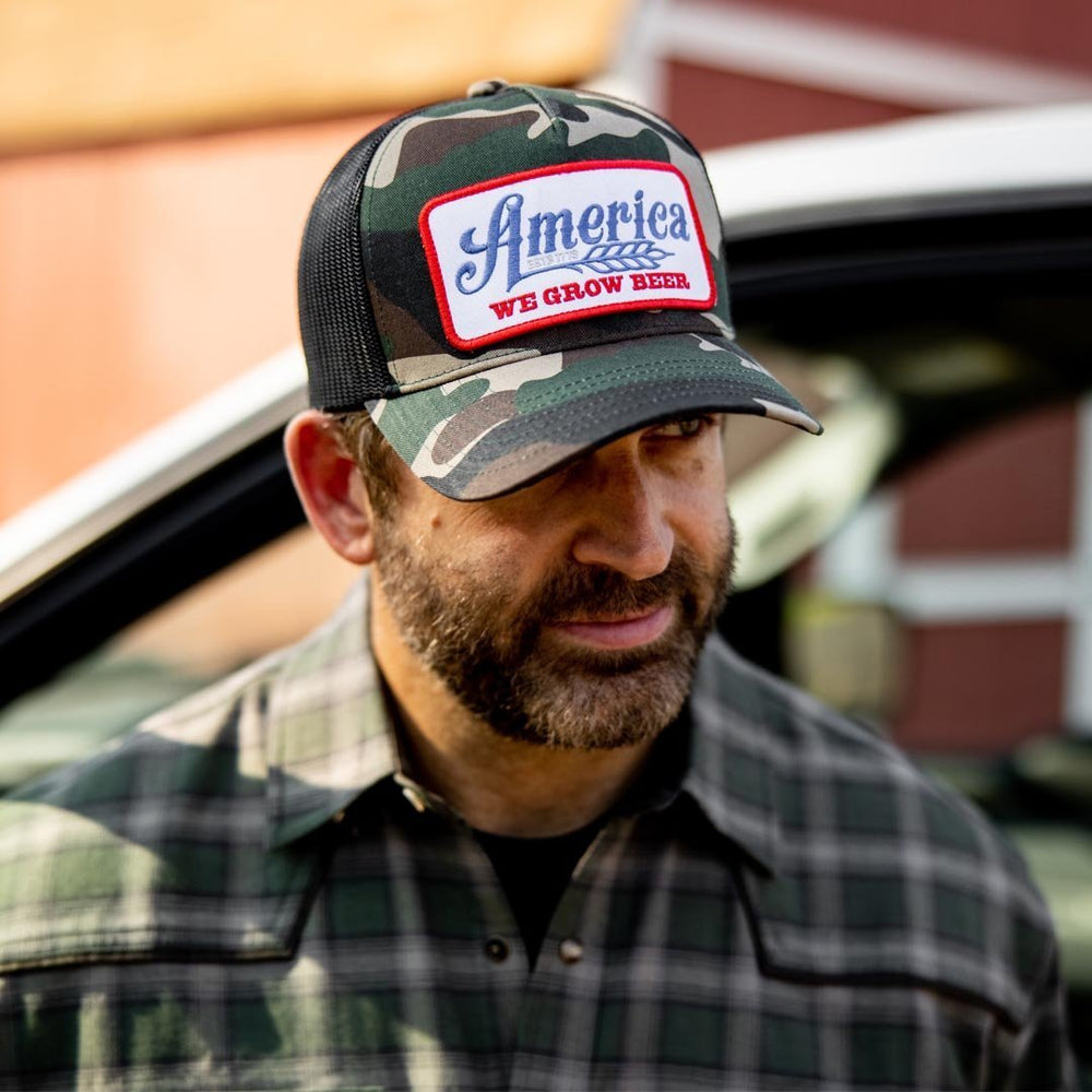 A man with a beard wears a heartland-inspired Rural Cloth "We Grow Beer Hat-Camo," a camouflage baseball cap adorned with a patch reading "America: We Grow Beer." Standing outdoors next to a vehicle, he is dressed in a green plaid shirt, embodying a tribute to hard-working Americans.