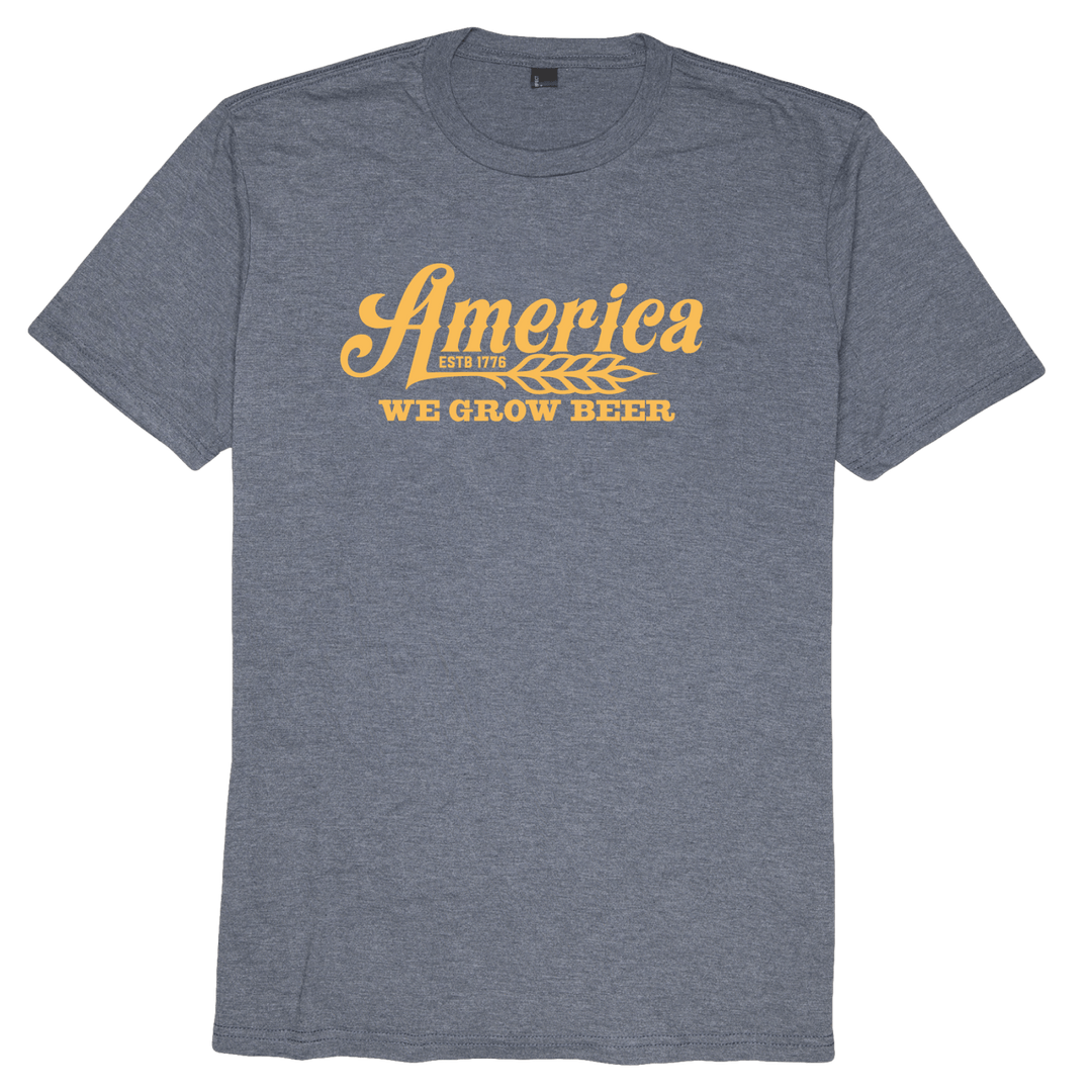 A navy frost t-shirt from Rural Cloth features a stylish yellow graphic design with the word "America" in a stylized font above the phrase "WE GROW BEER." There is also smaller text reading "ESTD 1776" and a wheat symbol below the main word.
