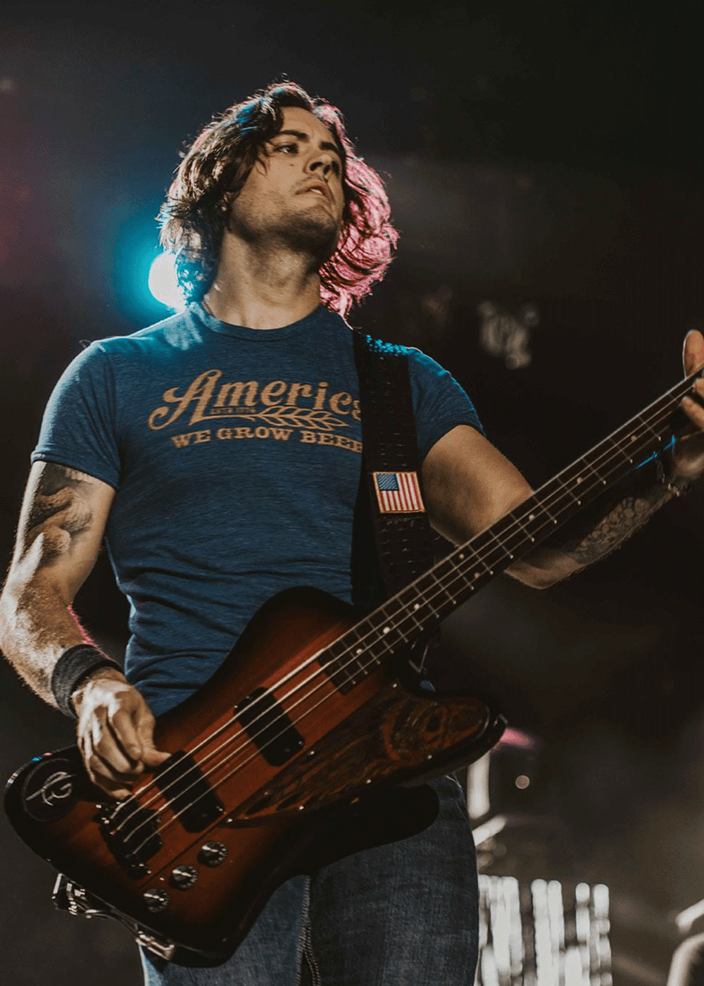 A person with shoulder-length hair stands on stage playing an electric bass guitar. They are wearing the We Grow Beer Tee - Royal from Rural Cloth, which is made from super soft combed ring-spun cotton and printed with "America: We Grow Beer." They also have on blue jeans and a wristband. Stage lights illuminate the background.