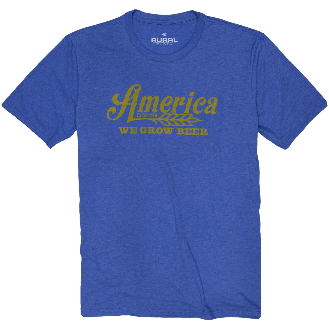 The We Grow Beer Tee - Royal by Rural Cloth is a blue T-shirt featuring the text "America like we grow beer" printed in mustard yellow across the chest, accompanied by an illustration of barley next to the word "America." Made from super soft combed ring-spun cotton, this shirt offers a modern athletic fit with the brand label "Rural" visible below the neckline.