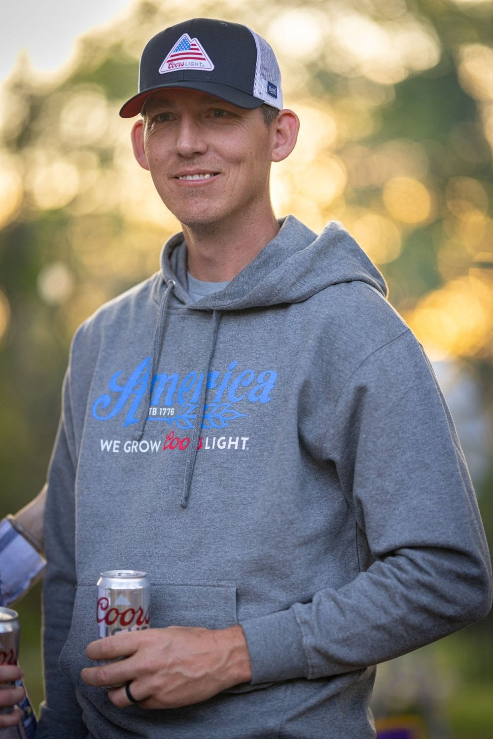 A person wearing the We Grow Coors Light Pullover in gray from Rural Cloth and a black-and-red baseball cap is holding a can of Coors Light, the iconic Silver Bullet. The background appears to be an outdoor setting with blurred greenery and sunset light.