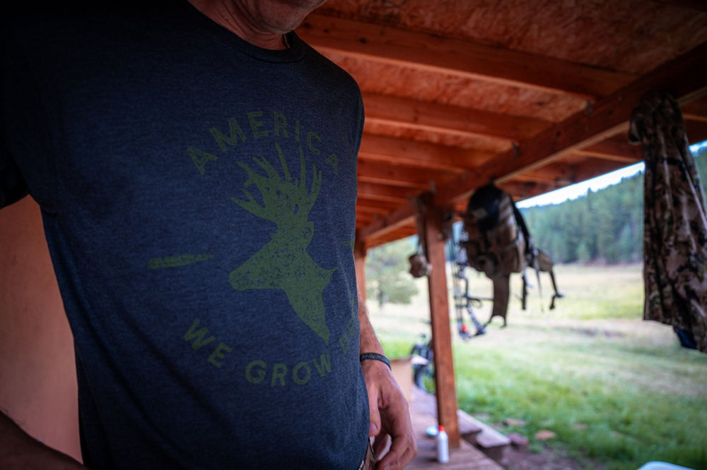 A person is standing outside under a wooden overhang, wearing a dark "We Grow Deer Tee-Black" by Rural Cloth, which features an illustrated deer head and the words "America We Grow." The background shows a grassy field, trees, and various gear hanging from the structure, reflecting a commitment to wildlife conservation.
