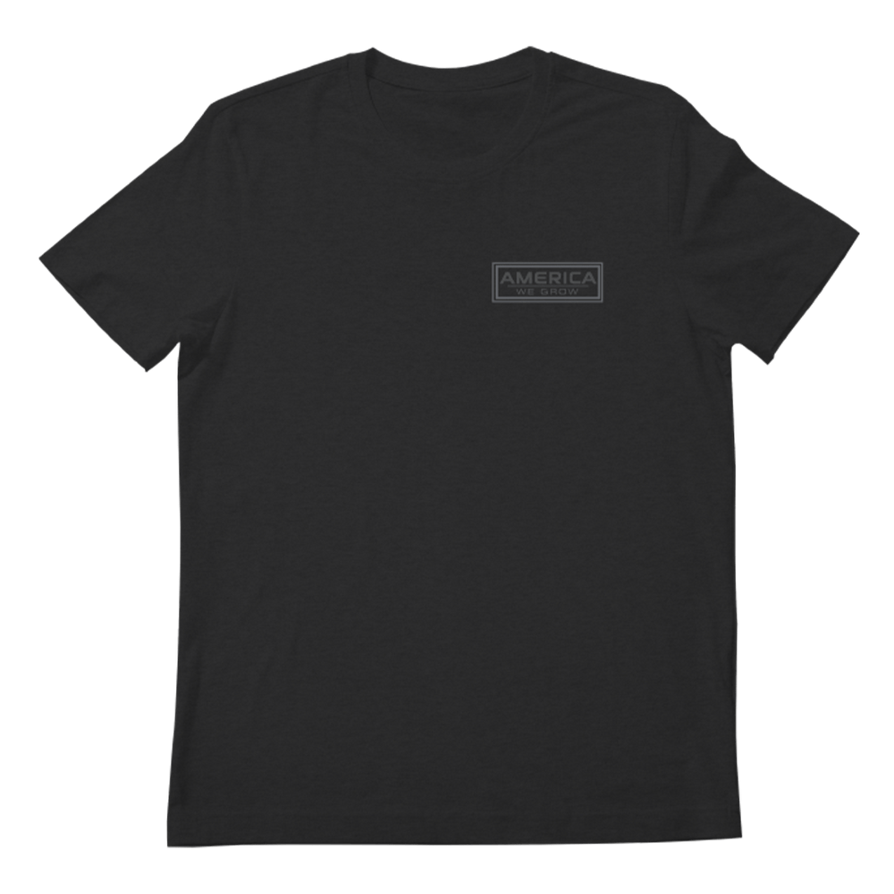 The We Grow Freedom Tee by Rural Cloth is a plain black short-sleeve T-shirt featuring a rectangular logo on the left chest area. The logo displays the word "AMERICA" in uppercase letters and subtly incorporates an American flag design. This patriotic tee boasts a minimalistic and monochromatic aesthetic, perfect for any casual occasion.