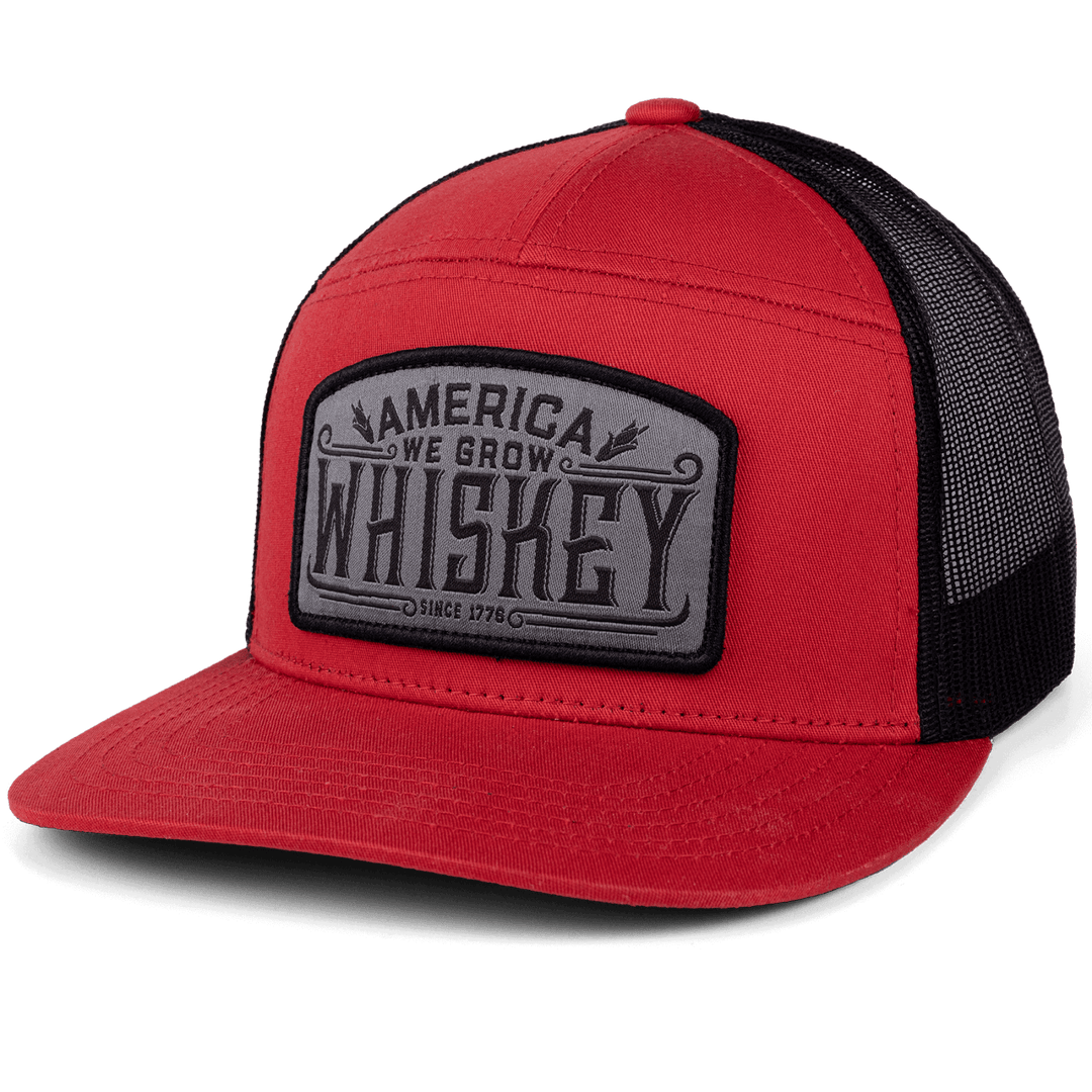 The Rural Cloth "We Grow Whiskey - 7-Panel" hat features a bold front patch with the phrase "America We Grow Whiskey Since 1776." The hat boasts a striking color scheme with the red front panel and bill, complemented by black mesh on the back and sides, paying homage to America's cherished spirit.