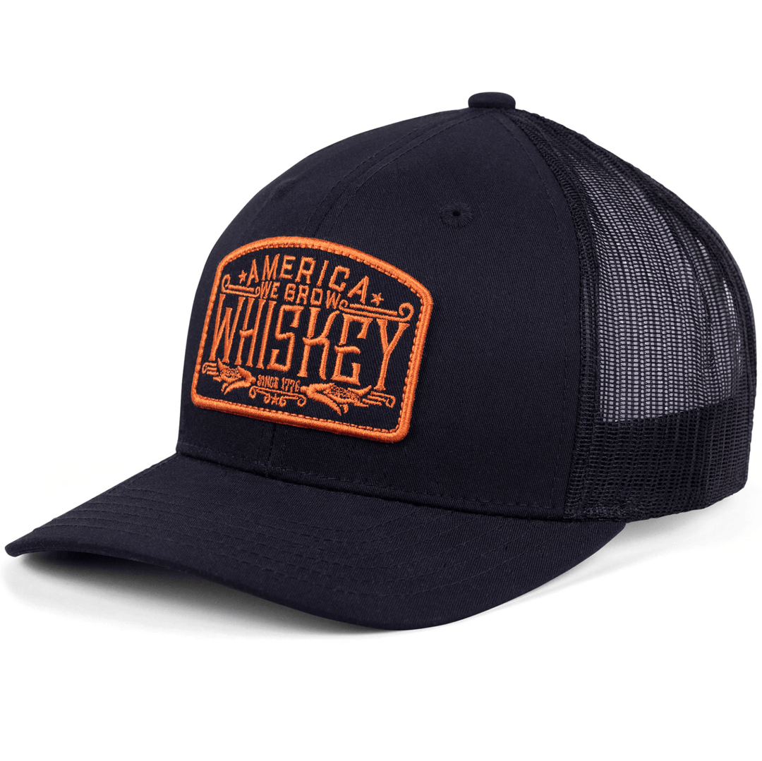 The "We Grow Whiskey Hat-Black/Orange" by Rural Cloth is a black mesh baseball cap featuring an orange embroidered patch on the front that reads "America We Grow Whiskey," surrounded by stars and arrows. It has a curved brim and an adjustable snapback closure for a perfect fit.
