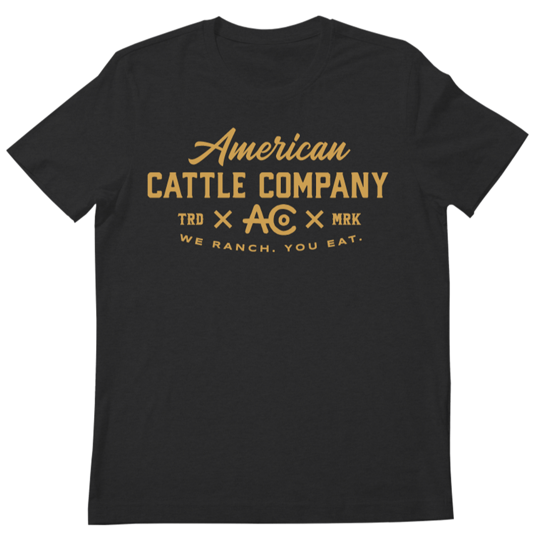 The We Ranch You Eat Tee-Heather Charcoal by Rural Cloth is a black T-shirt with the gold text "American Cattle Co" prominently displayed on the front. Below it, you'll find smaller texts and symbols including "TRD," "AC" in a circle, and "MRK." At the bottom, the slogan "We Ranch. You Eat." is printed. This stylish tee pays tribute to American cattle ranchers and showcases their heritage with flair.