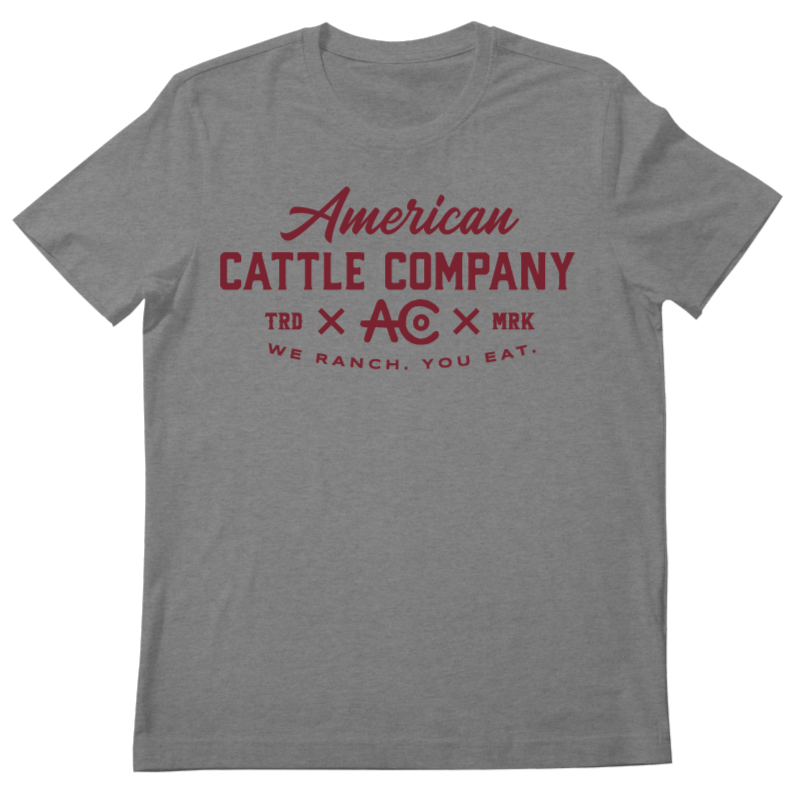 The We Ranch You Eat Tee-Heather Gray by Rural Cloth is a grey T-shirt that showcases "American Cattle Co" in red, with "TRD AC MRK" and "We Ranch You Eat" also in red beneath, all centered on the shirt.