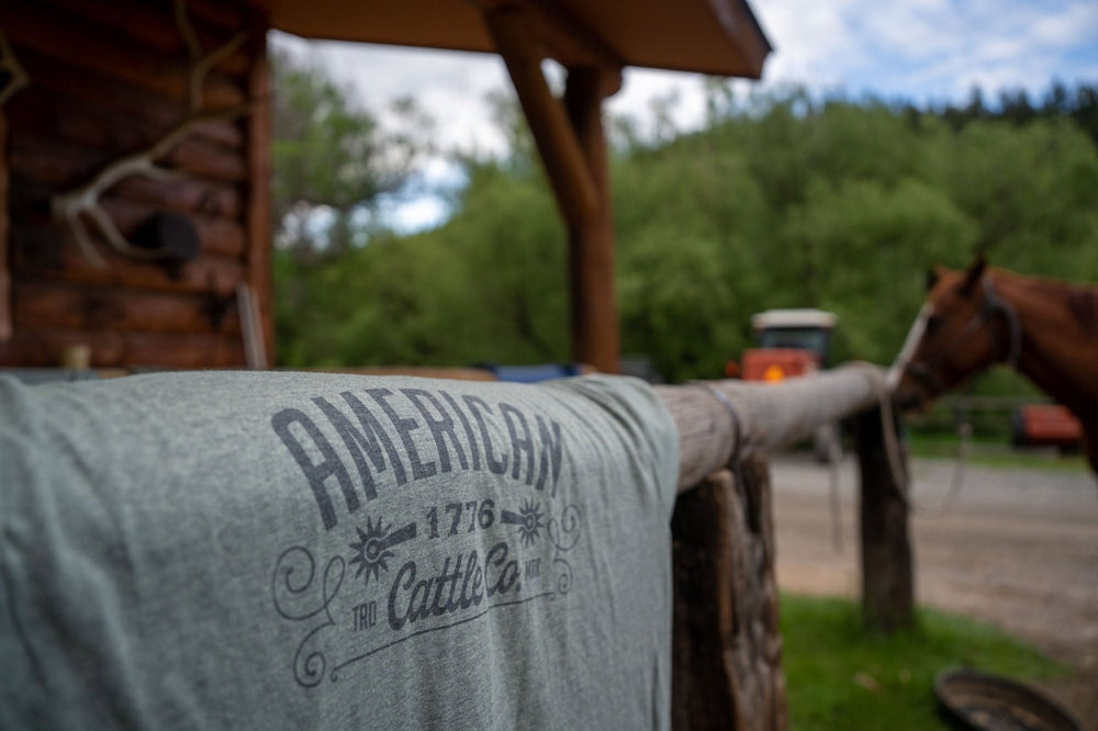 A gray Western Tee from Rural Cloth, printed with "American 1776 The Cattle Co.," is draped over a wooden railing. Comfortable and durable, it perfectly captures the spirit of the American cattle industry. In the background, a horse stands near a wooden cabin in a forested area, with a tractor visible in the distance under a cloudy sky.